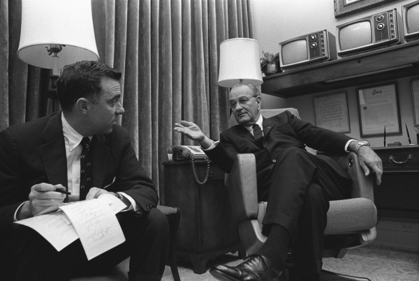 Black-and-white photograph of two men in suits seated in armchairs. One holds a notepad and pen; the other gestures while speaking, with television sets and framed documents visible behind him