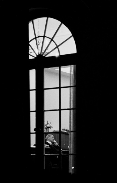 Black-and-white photograph of a man seated at a desk, seen through a tall arched window, holding a telephone receiver to his ear