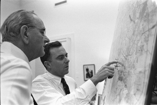Black-and-white photograph of two men in white dress shirts studying a large wall map, one pointing to a location marked with pins