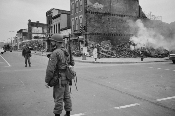 Black-and-white photograph of an armed soldier in helmet and field gear standing in an intersection beside piles of rubble and a smoldering brick building, with a second soldier and a civilian visible in the background
