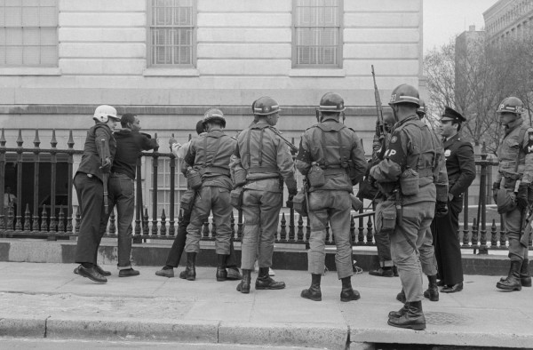 Black-and-white photograph of helmeted soldiers and military police, armed with rifles, standing on a sidewalk before an iron fence and neoclassical building