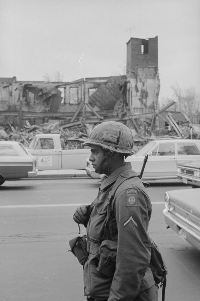 Black-and-white photograph of an armed soldier in helmet and combat gear standing on a street in front of a gutted, collapsed building and parked cars