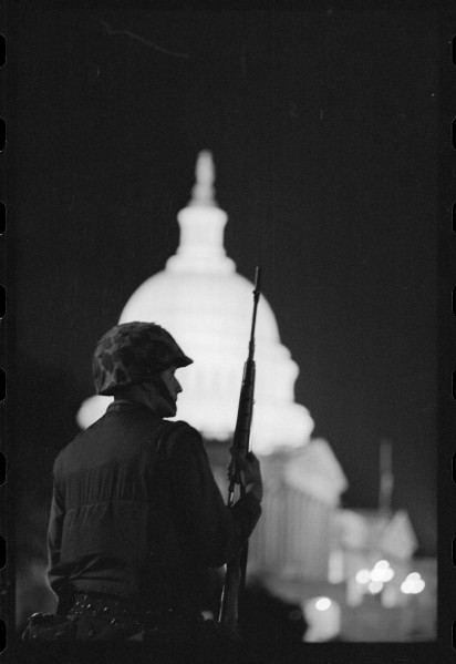Black-and-white photograph of an armed, helmeted soldier in profile at night, with the illuminated U.S. Capitol dome glowing in the background