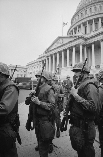 Black-and-white photograph of armed soldiers in helmets and fatigues standing before the U.S. Capitol, its dome visible with a flag at half-staff