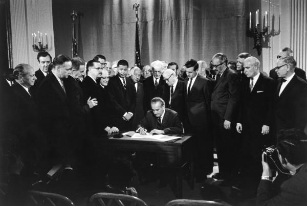 Black-and-white photograph of a man seated at a desk signing a document, surrounded by a large group of men in suits. An American flag and candelabras are visible in the background