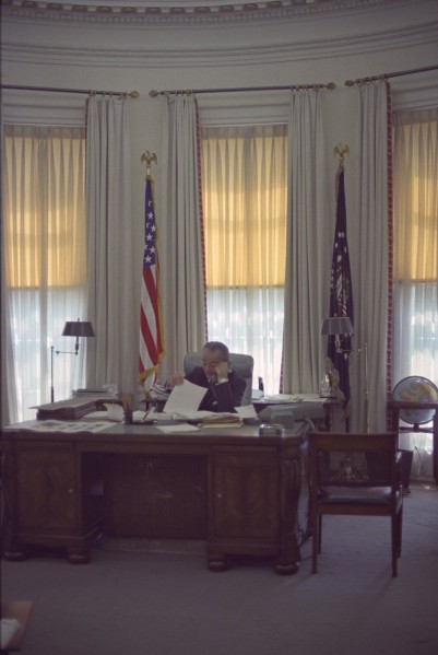 Color photograph of a man seated at a large wooden desk, talking on a telephone and holding papers, flanked by the U.S. flag and a dark ceremonial flag