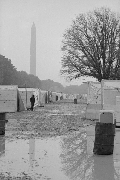 Black-and-white photograph of a muddy encampment of plywood-and-canvas shelters with the Washington Monument rising in the misty background