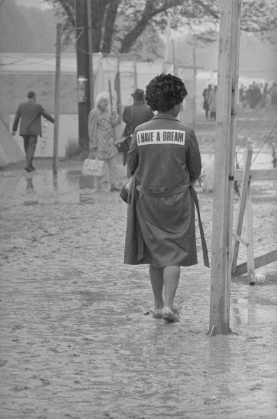 Black-and-white photograph of a woman walking barefoot through deep mud, her coat bearing a patch reading "I HAVE A DREAM," with tents and people visible behind her