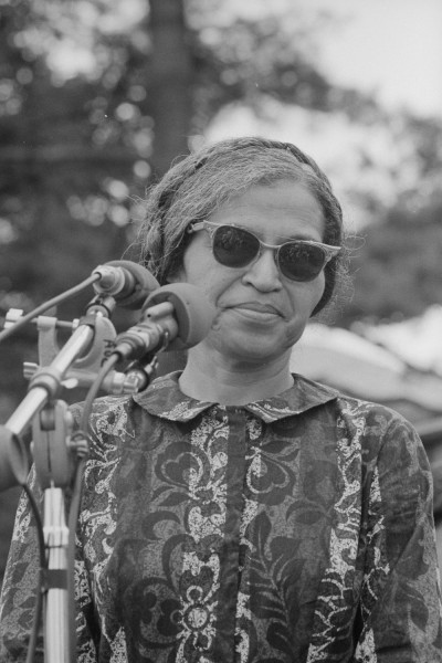 Black-and-white photograph of a woman wearing sunglasses and a patterned blouse standing at a cluster of microphones outdoors, trees visible behind her
