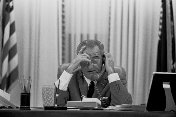 Black-and-white photograph of an older man in a suit seated at a large desk, holding a telephone receiver to his ear with both hands raised to his head