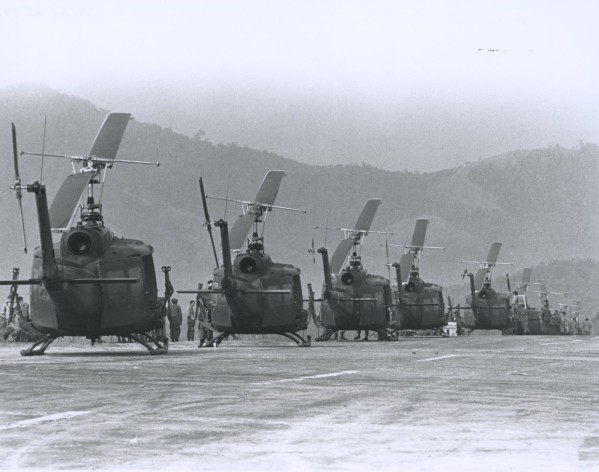 Black-and-white photograph of a row of military helicopters parked on a tarmac, with figures standing nearby and forested hills in the background