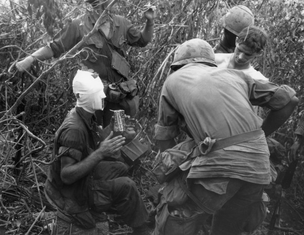 Black-and-white photograph of several soldiers in combat gear amid dense brush; one man is seated with his head and face heavily bandaged, holding a Cracker Jack box