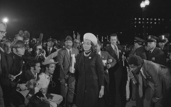 Black-and-white photograph of a woman in a white head covering holding a candle at night, surrounded by a dense crowd of marchers, press photographers, and police officers