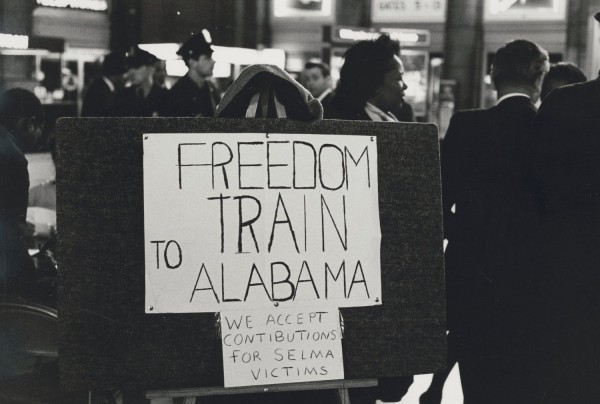 Black-and-white photograph of a handmade sign on an easel reading "FREEDOM TRAIN TO ALABAMA" with a smaller note reading "WE ACCEPT CONTIBUTIONS FOR SELMA VICTIMS," in a busy indoor public space with figures visible in the background