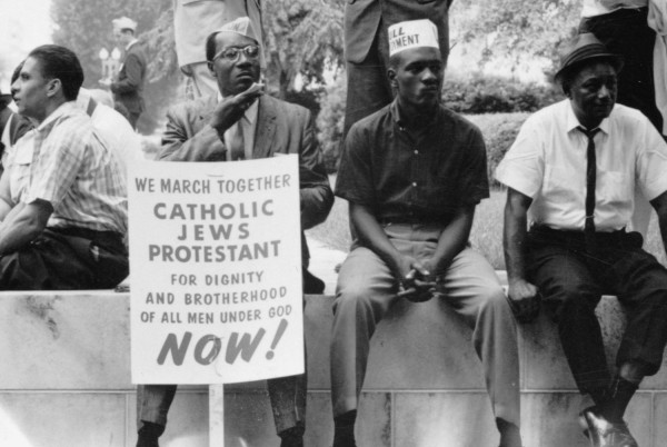 Black-and-white photograph of several men seated on a low stone wall, one holding a sign reading "We March Together — Catholic, Jews, Protestant — For Dignity and Brotherhood of All Men Under God — Now!"