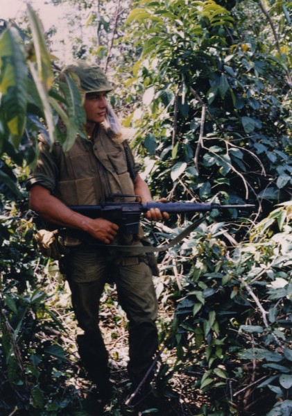 Color photographic print of a soldier in olive fatigues and flak vest holding a rifle with bayonet, standing among dense jungle foliage