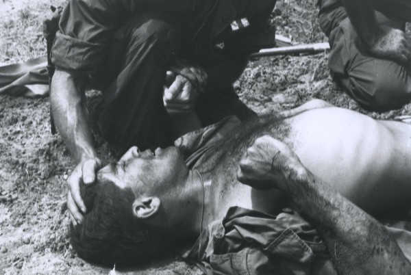 Black-and-white photograph of a shirtless man lying on the ground, eyes closed, while another person in military clothing kneels over him