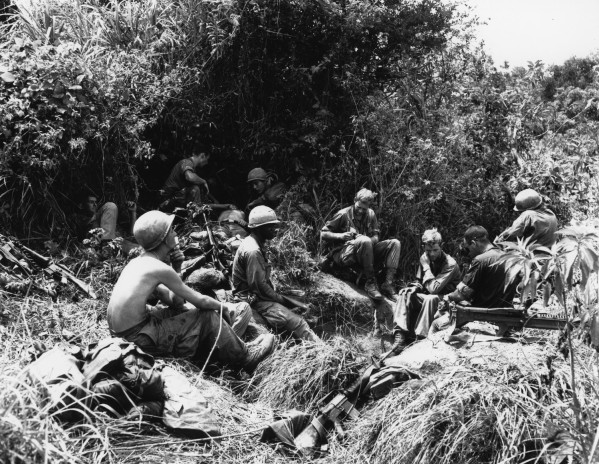 Black-and-white photograph of a group of soldiers in combat gear resting on a hillside amid dense jungle vegetation, rifles visible nearby