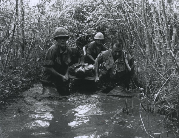 Black-and-white photograph of three soldiers in helmets wading through a flooded swamp, carrying a fourth soldier on a stretcher through dense vegetation