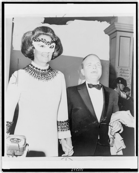 Black-and-white photo of a woman in a jeweled mask and white gown holding hands with a shorter man in a tuxedo, smiling indoors