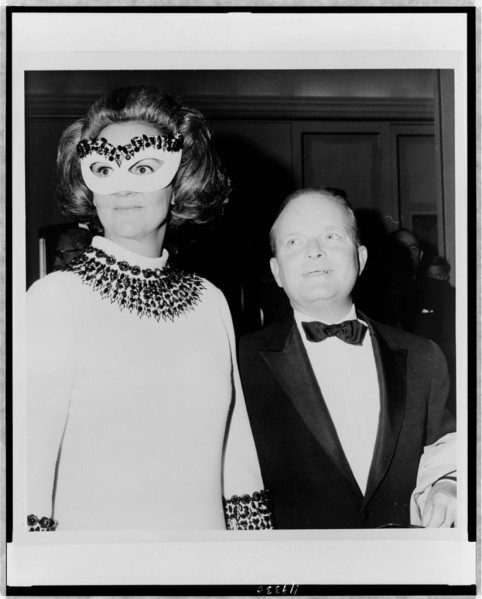 Black-and-white photograph of a woman in a jeweled eye mask and embellished white dress standing beside a heavyset man in a tuxedo and bow tie