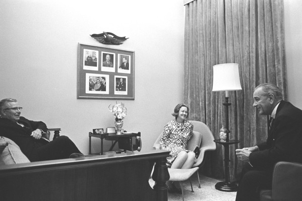 Black-and-white photograph of three people seated in armchairs in an office, with a framed photo collage and decorative eagle on the wall behind them