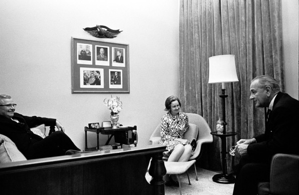 Black-and-white photograph of three people seated in a formal interior, conversing; a framed collage of portraits and a decorative eagle hang on the wall behind them