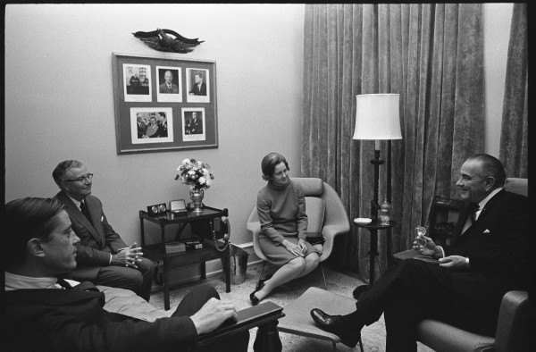 Black-and-white photograph of four people seated in armchairs in a wood-paneled room; a framed collage of portraits hangs on the wall behind them