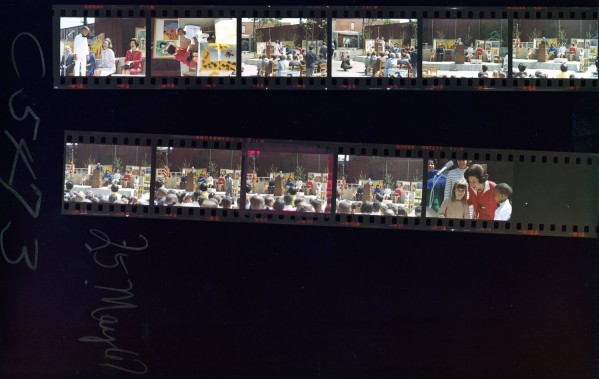 Color contact sheet with two strips of film frames showing an outdoor dedication ceremony with a crowd, a speaker's podium, colorful artwork displays, and groups of adults and children