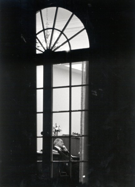 Black-and-white photograph shot through a tall arched White House window; a man seated at a desk inside holds a telephone receiver to his ear