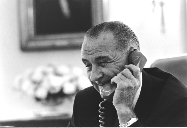 Black-and-white photograph of an older man in a dark suit holding a telephone handset to his ear, smiling slightly, seated at a desk with a framed painting visible behind him