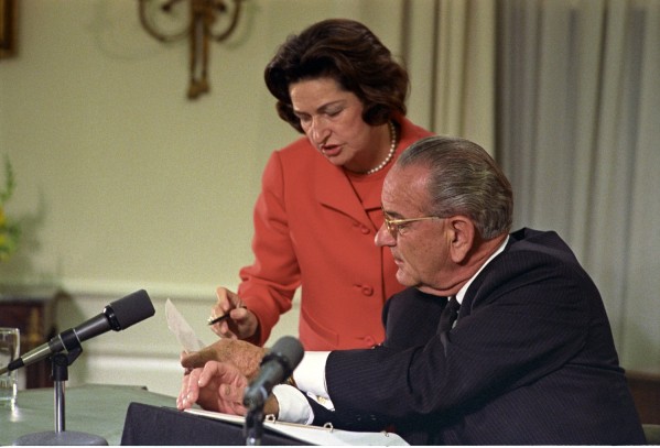 Color photograph of a woman in a red jacket standing behind a seated man in a dark suit at a table with microphones, both looking down at papers