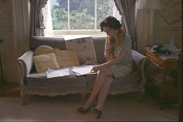 Color photograph of a woman seated on a sofa, holding a handheld microphone and reviewing handwritten notes and newspapers; a reel-to-reel recorder sits on a side table nearby