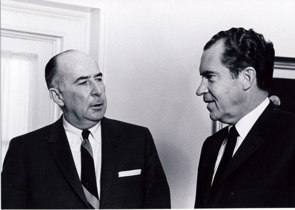 Black-and-white photograph of two men in dark suits and ties facing each other in conversation against a light-colored wall near a doorway
