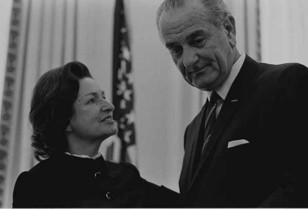 Black-and-white photograph of a woman smiling up at a taller man in a dark suit and tie, an American flag visible behind them