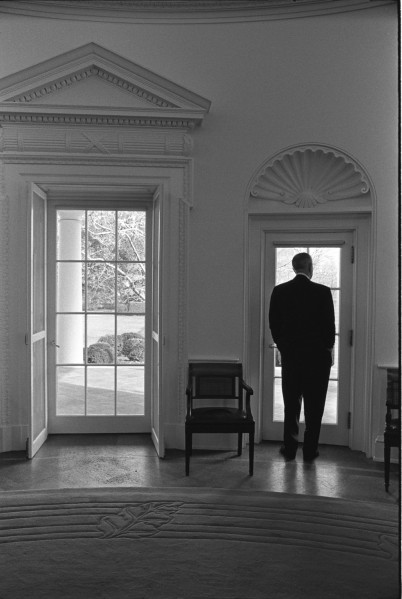 Black-and-white photograph of a man in a dark suit standing with his back to the camera, looking out a glass-paned door in an ornately trimmed room