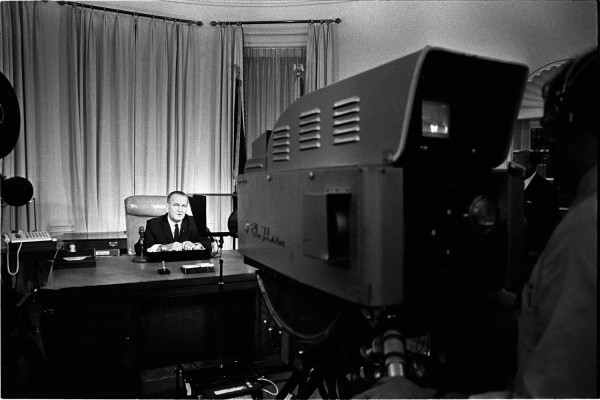 Black-and-white photograph of a man in a dark suit seated at a desk before drawn curtains, with a large television broadcast camera in the foreground