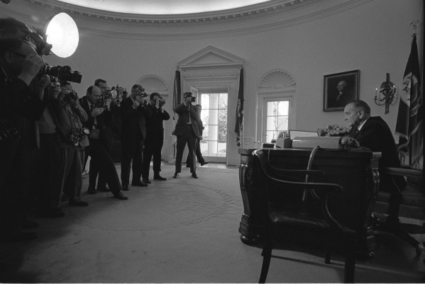 Black-and-white photograph of a man seated at a large desk in a round, ornately molded room while a crowd of photographers with cameras and lights aim at him from the left