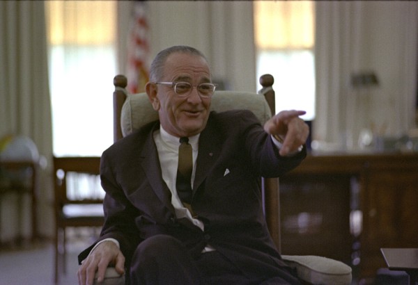 Color photograph of a man in a dark suit and glasses seated in a cushioned rocking chair, pointing outward, with a desk and American flag visible behind him