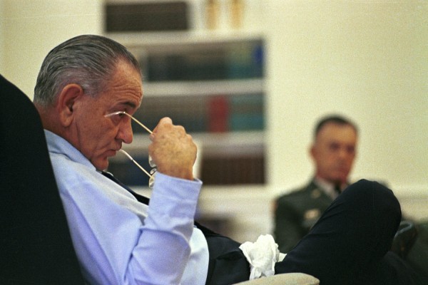 Color photograph of a man in a light blue shirt seated in a chair, holding eyeglasses to his nose; a uniformed military officer sits blurred in the background