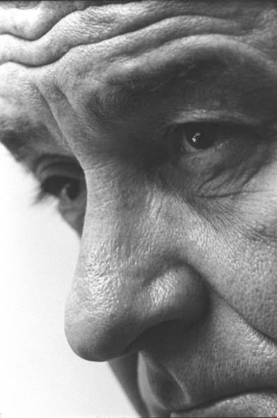 Black-and-white close-up photograph of an older man's face, showing deeply lined forehead, nose, and one eye in sharp focus against a light background