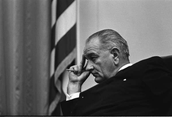 Black-and-white photograph of a man in a dark suit seen in close profile, resting his temple on raised fingers while holding a pencil, an American flag visible behind him