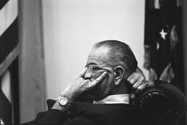 Black-and-white photograph of a man in profile, wearing glasses and a suit, hands raised to his face, seated in a leather chair with a U.S. flag behind him
