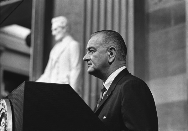 Black-and-white photograph of a man in a dark suit speaking at a presidential podium, with a large seated marble statue visible behind him
