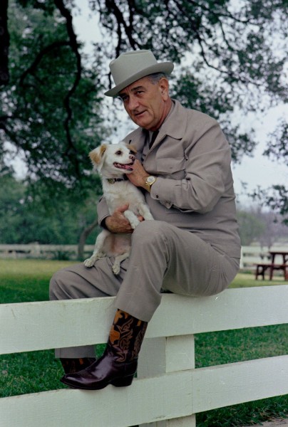 Color photograph of a man in a gray suit, wide-brimmed hat, and ornate cowboy boots seated on a white fence, holding a small white dog