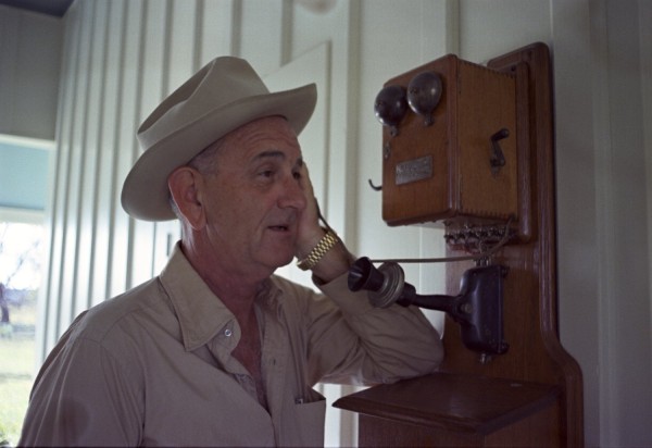 Color photograph of a man in a white cowboy hat and khaki shirt holding the handset of a wall-mounted antique wooden telephone