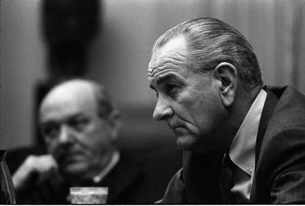 Black-and-white photograph of a man in profile seated at a table, looking downward, with a second man visible out of focus in the background