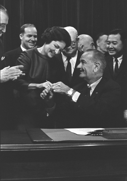 Black-and-white photograph of a seated man handing a pen to a smiling woman leaning toward him, surrounded by a crowd of smiling onlookers