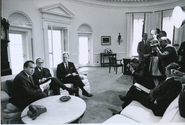 Black-and-white photograph of three men seated in armchairs in a large, formally furnished oval room while several photographers and cameramen document the meeting