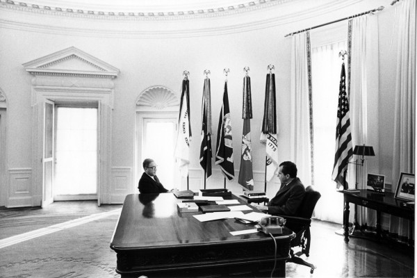 Black-and-white photograph of two men seated across a large wooden desk in a curved, formal office with multiple flags displayed behind them